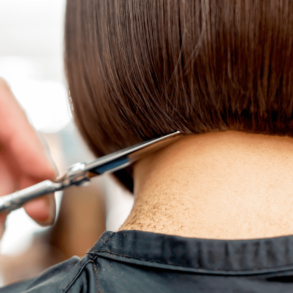 A close-up view of a precise haircut in progress, showing a stylist using professional scissors to trim the nape of a woman's neatly shaped bob haircut. The client wears a black salon cape, and freshly cut hair clippings can be seen on the neck, highlighting attention to detail and expert technique.